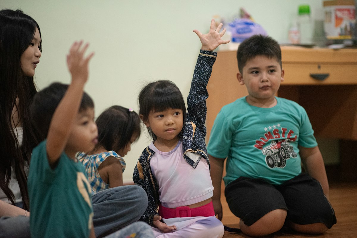 Grupo de niños sentados en el piso levantando la mano para participar junto a su teacher